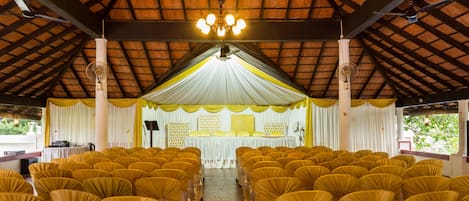 Un interno con file di sedie, un palco con uno sfondo bianco e un soffitto con decorazioni in legno.