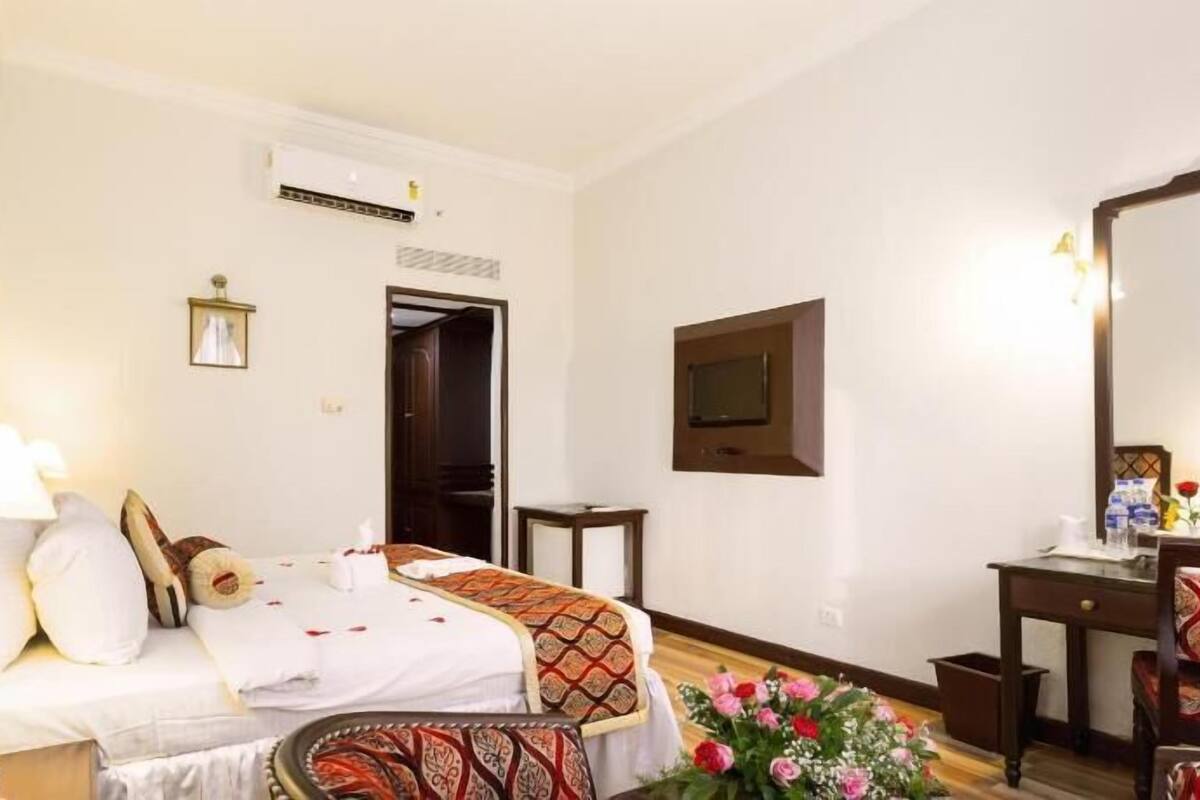 Habitación de hotel con cama, mesita de noche, lámpara, televisión y una pequeña mesa con flores.