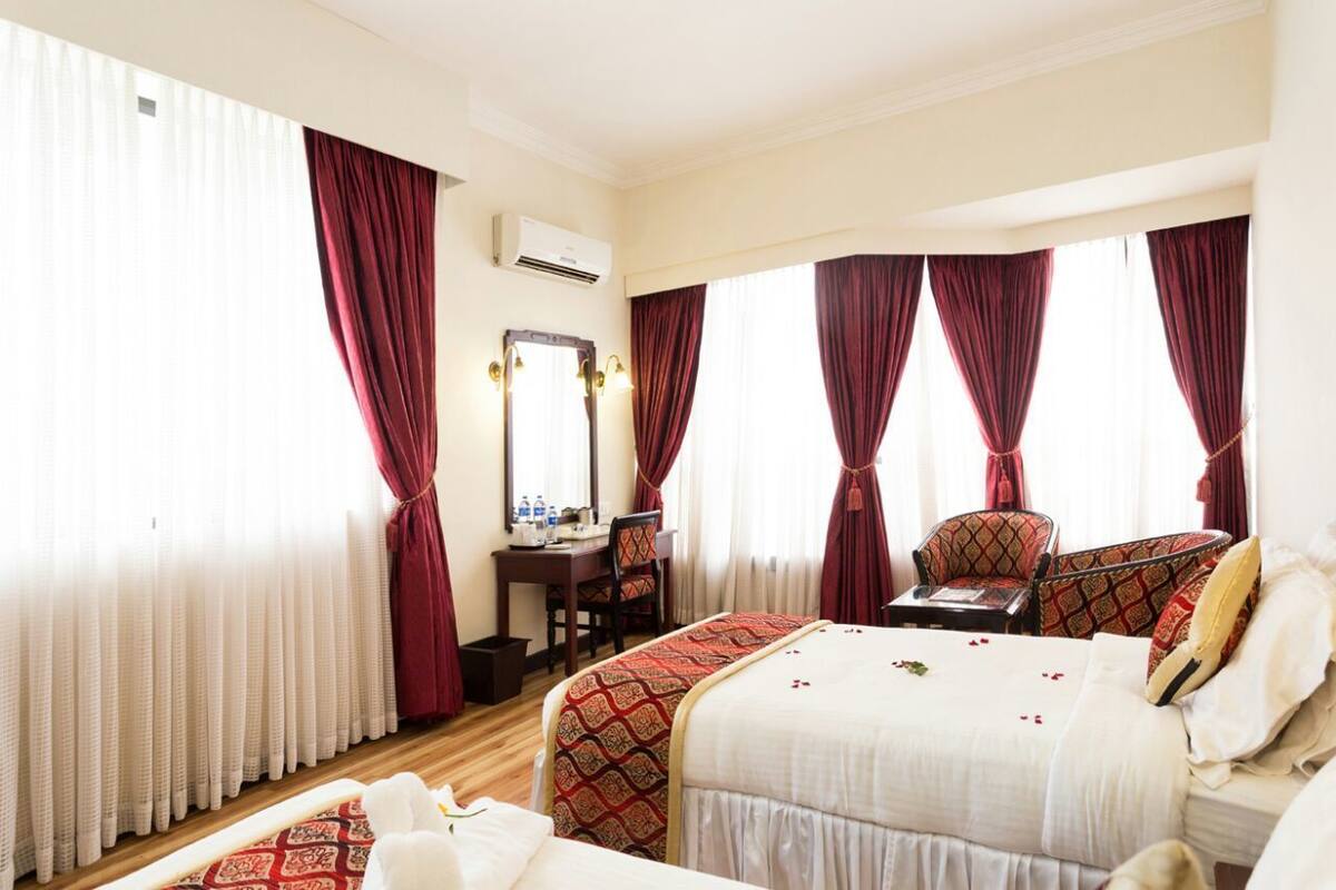 Habitación de hotel con una cama grande, un escritorio con silla, un espejo y dos ventanas con cortinas rojas.