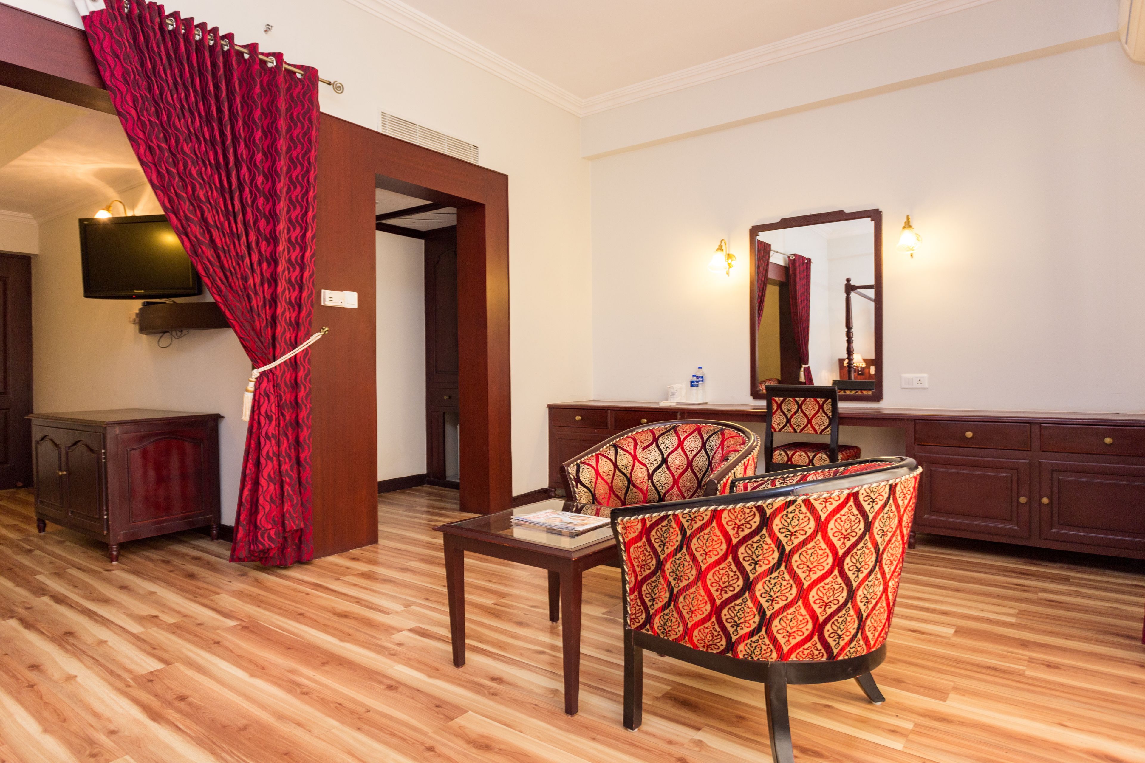 Quarto de hotel com piso de madeira, cortina vermelha, televisão, espelho e escrivaninha com cadeira.