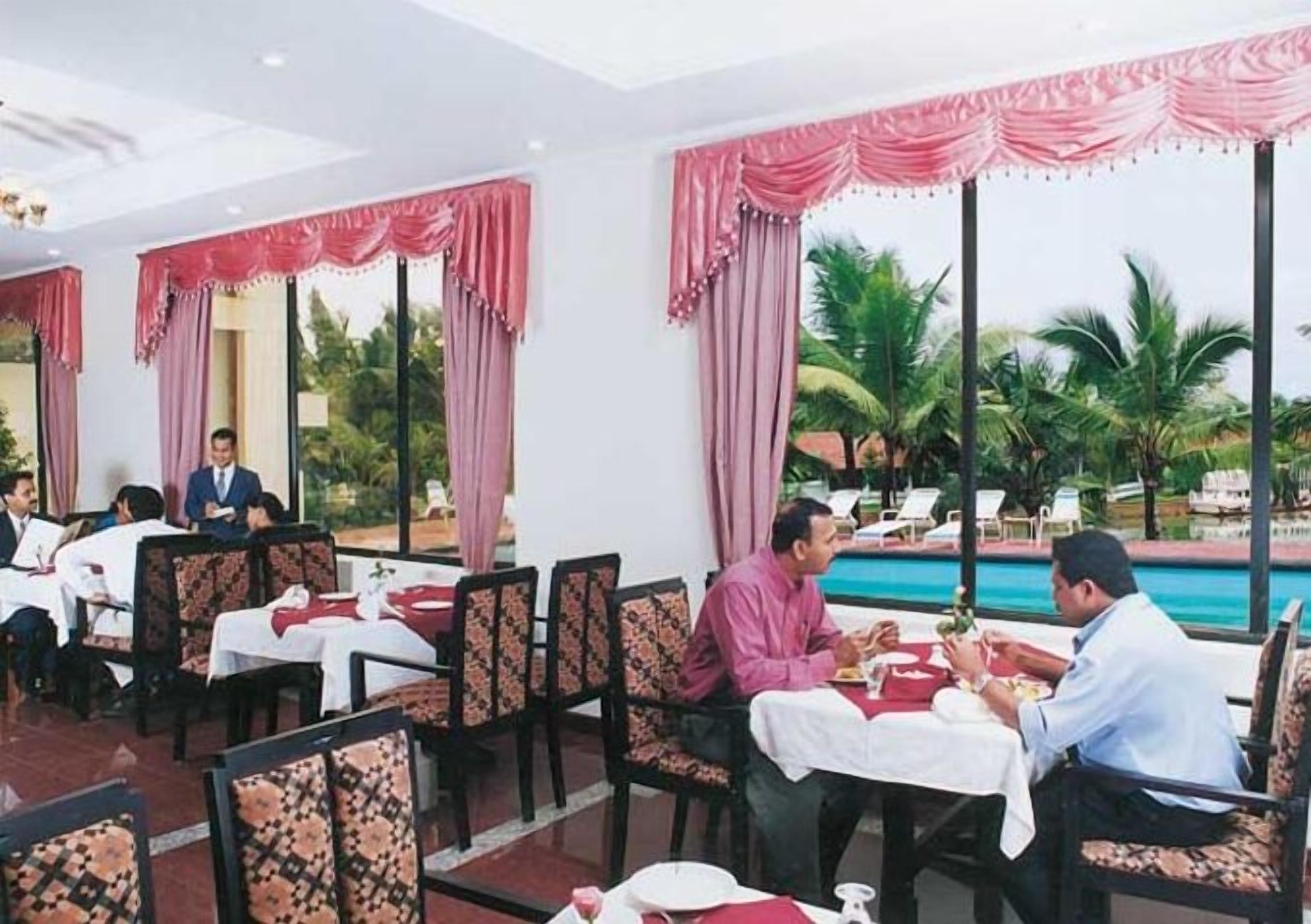 A restaurant with tables set for dining, large windows with curtains, and a view of palm trees and a pool.