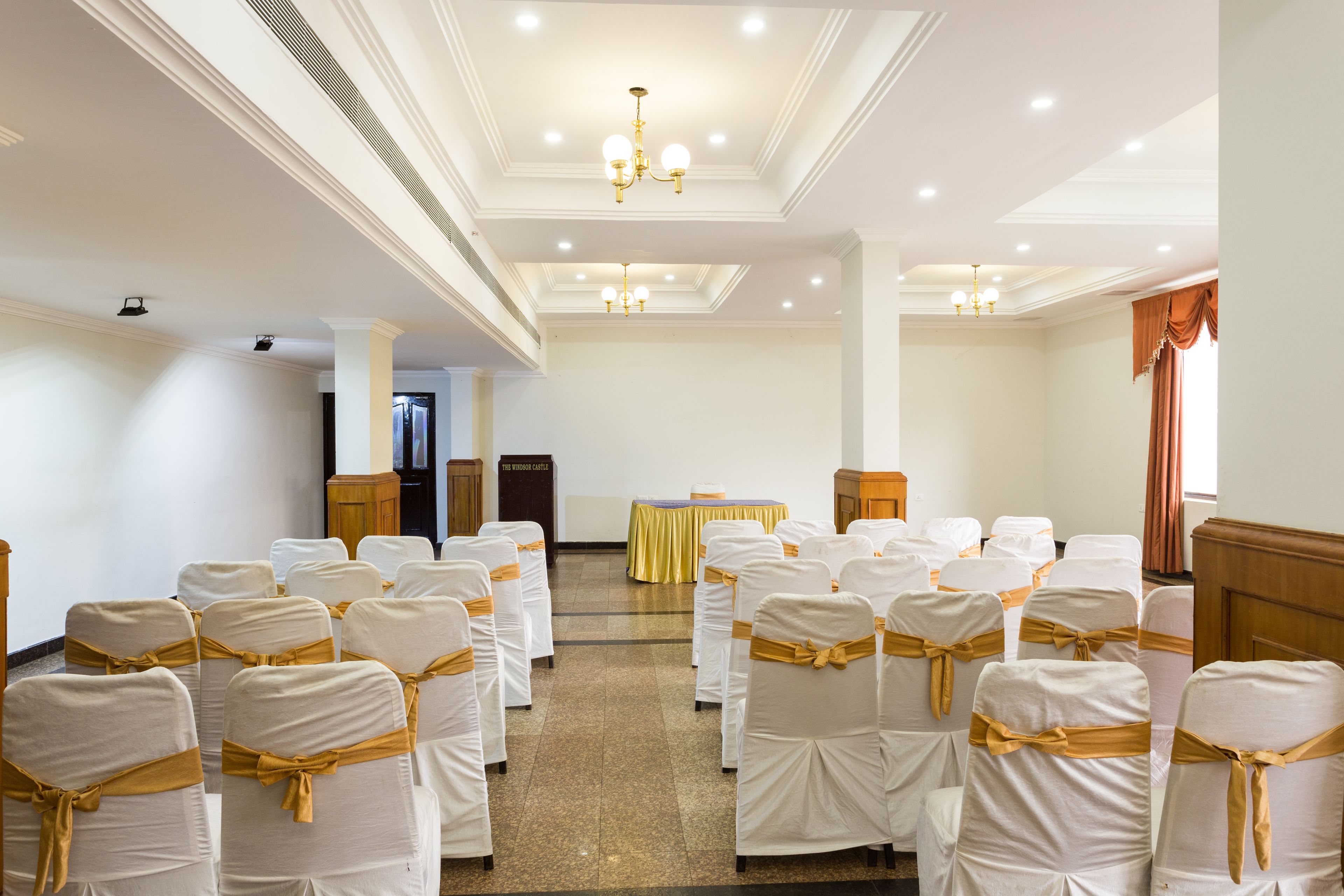 Un salón de conferencias con filas de sillas blancas adornadas con cintas doradas, un atril y una mesa con mantel amarillo.