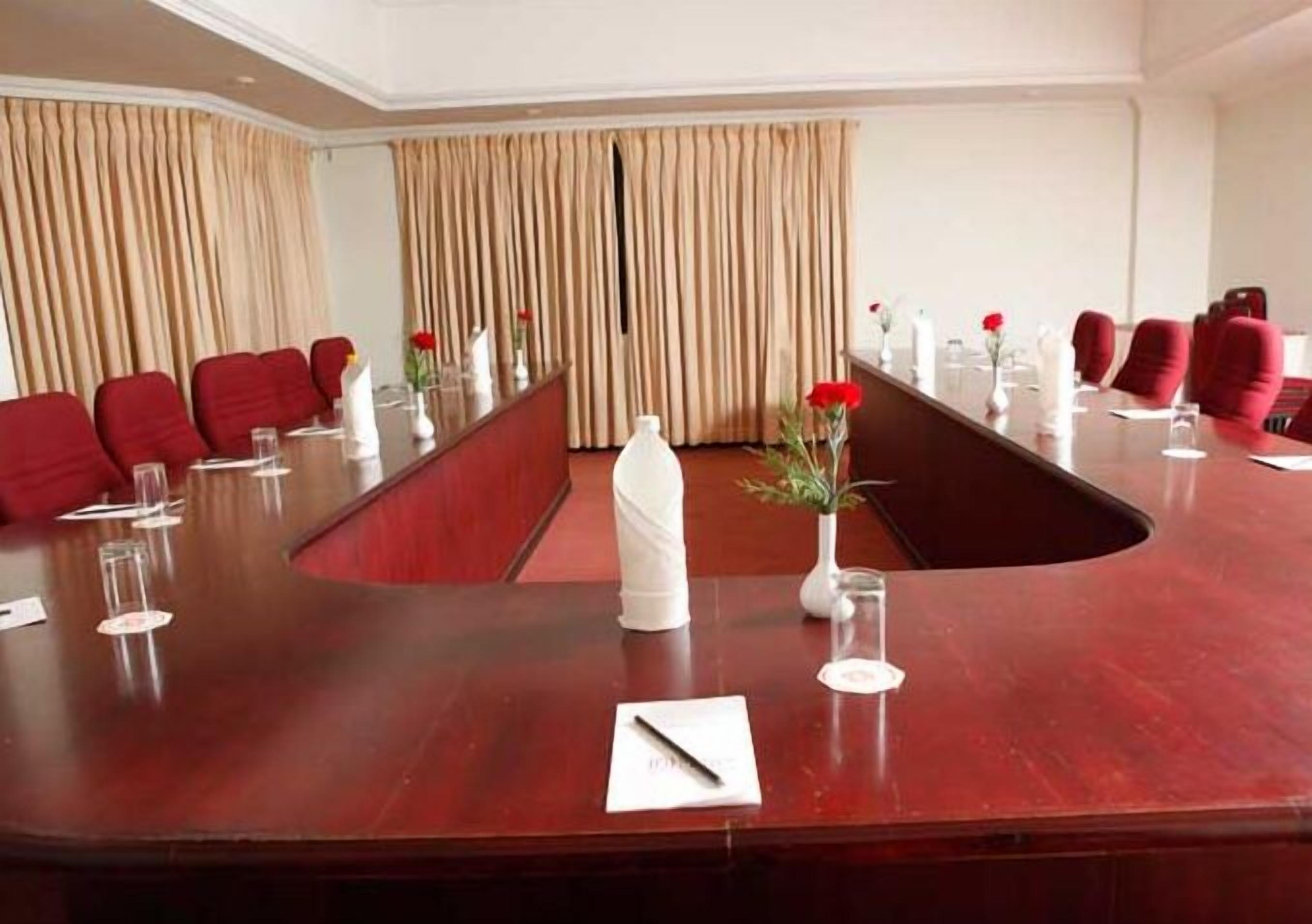 A conference room with a long table, red chairs, and a white vase with red flowers.