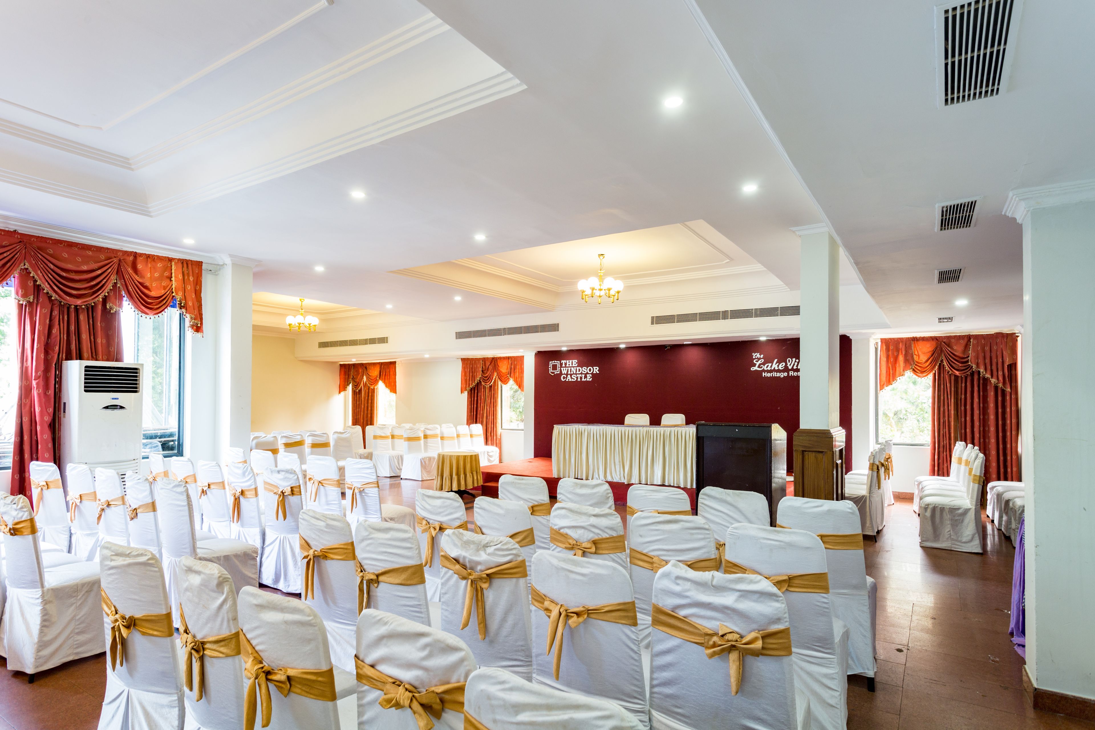 Un salón de banquetes con filas de sillas blancas con cintas doradas, un escenario con fondo rojo y una araña.