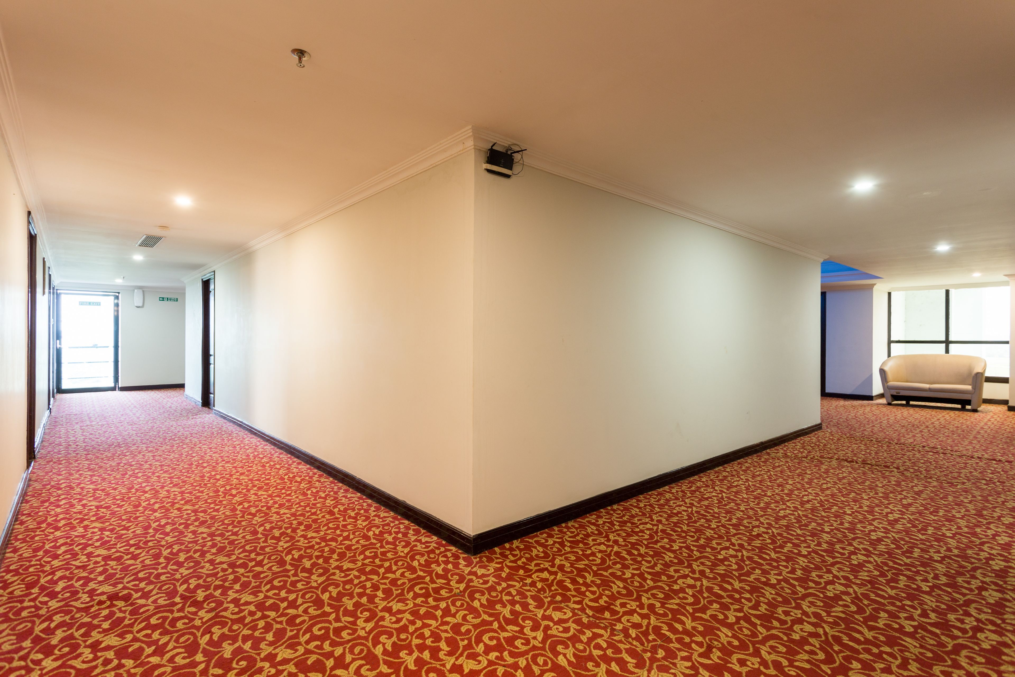 A hotel corridor with a red carpet, white walls, and a beige sofa in the corner.