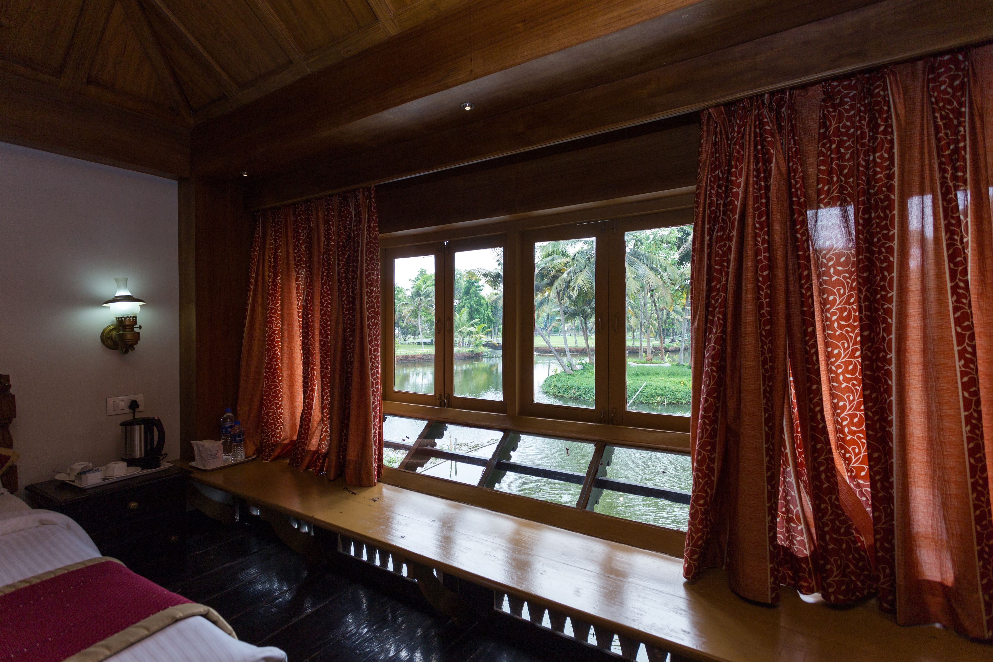 A room with a wooden bench along a window with patterned curtains, a bed, a nightstand with a lamp, and a view of greenery outside.