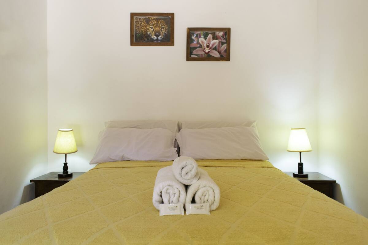 Cama bem arrumada com colcha amarela, dois travesseiros e duas toalhas enroladas com etiqueta. Há duas luminárias de mesa e duas fotos emolduradas na parede.