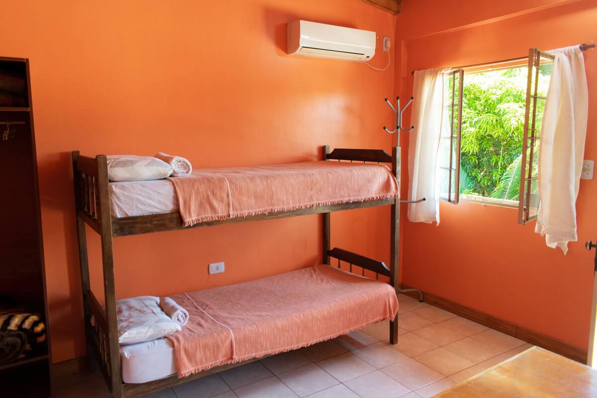 Quarto com beliche, ar condicionado, janela com cortinas e vista para o verde lá fora.