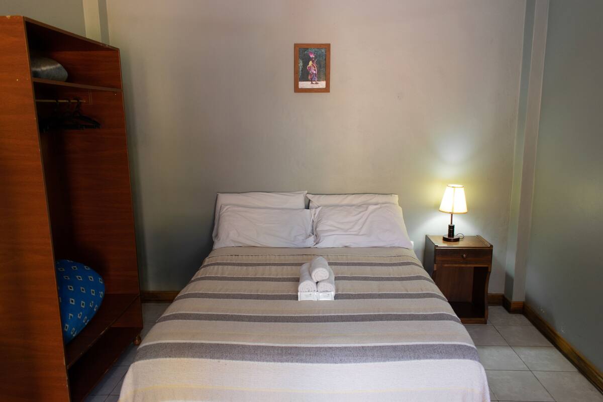 Quarto de hotel com cama, mesa de cabeceira com luminária, guarda-roupa de madeira e quadro na parede.