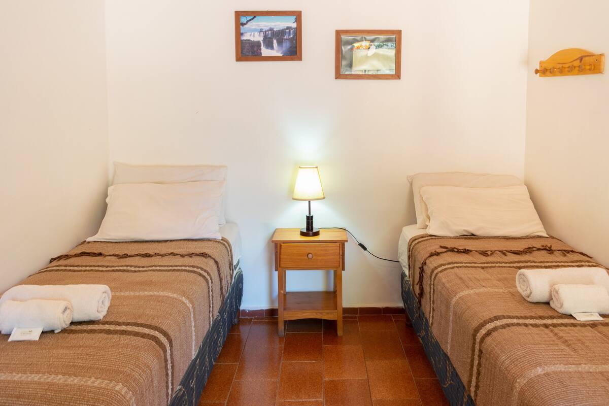 Quarto duplo com duas camas, mesa de cabeceira com abajur e duas quadros na parede.