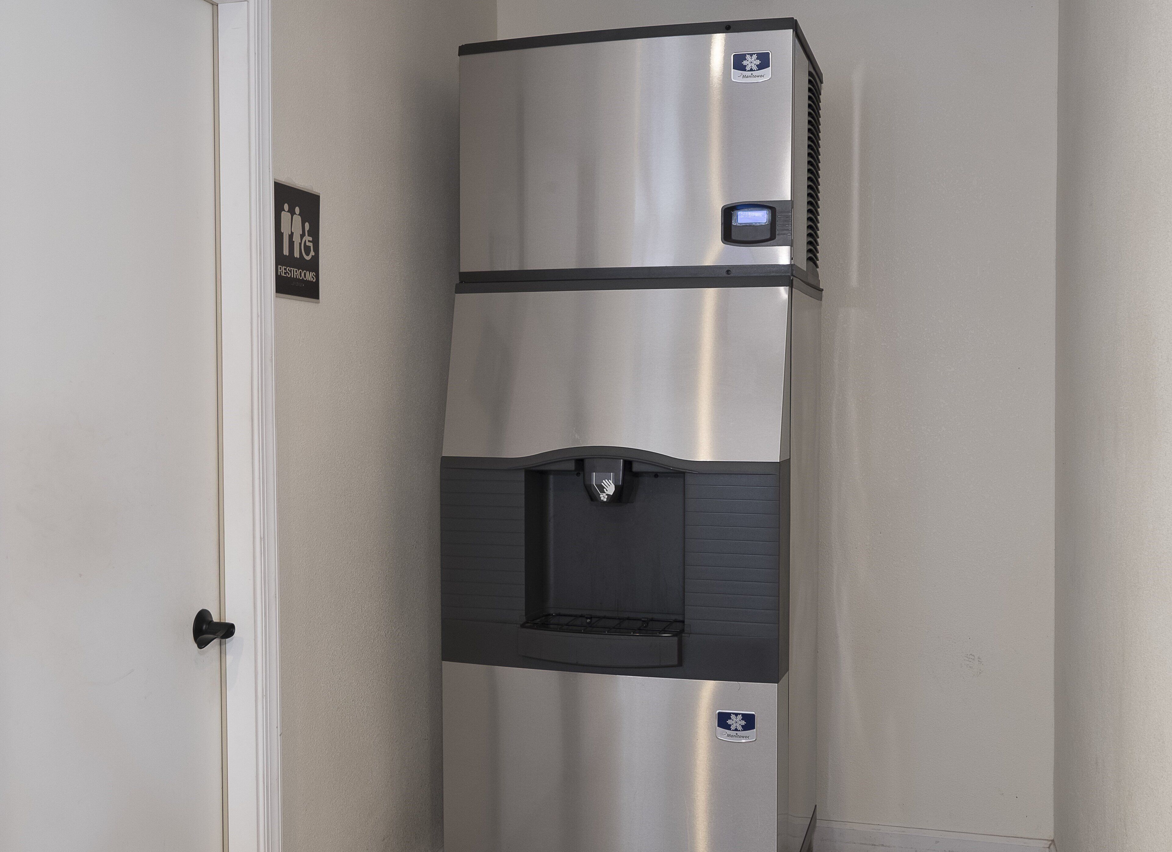 A stainless steel refrigerator with a water dispenser and a digital display, located in a hallway with a door and a sign indicating restrooms.