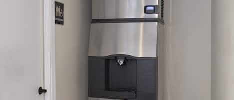 A stainless steel refrigerator with a water dispenser and a digital display, located in a hallway with a door and a sign indicating restrooms.