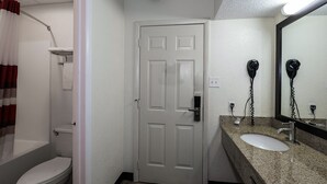 A bathroom with a white door, a toilet, a shower, and a sink with a mirror. - Red Roof Inn Lancaster - Strasburg (Lancaster)
