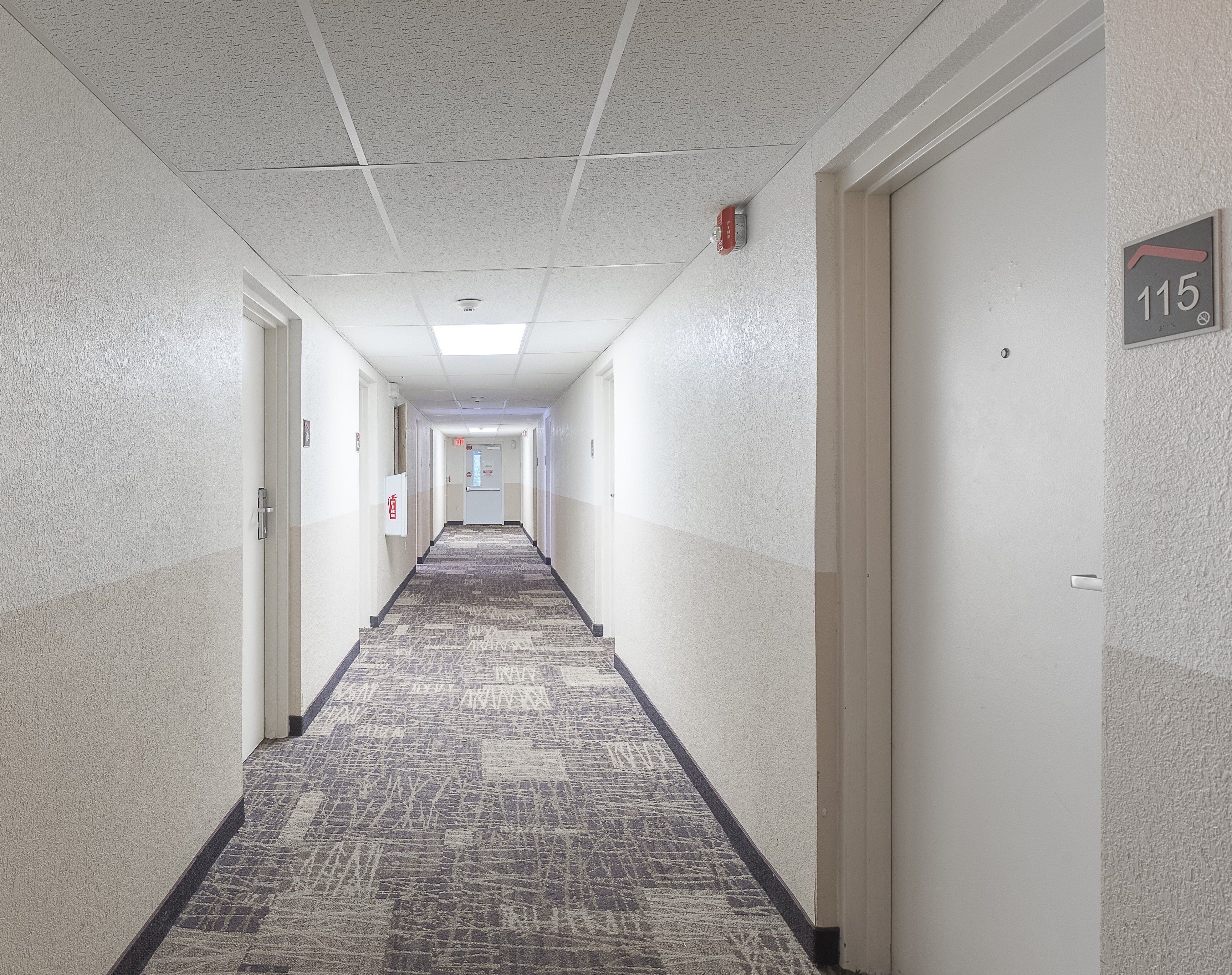 A hotel hallway with carpeted floors, white walls, and doors numbered 115.