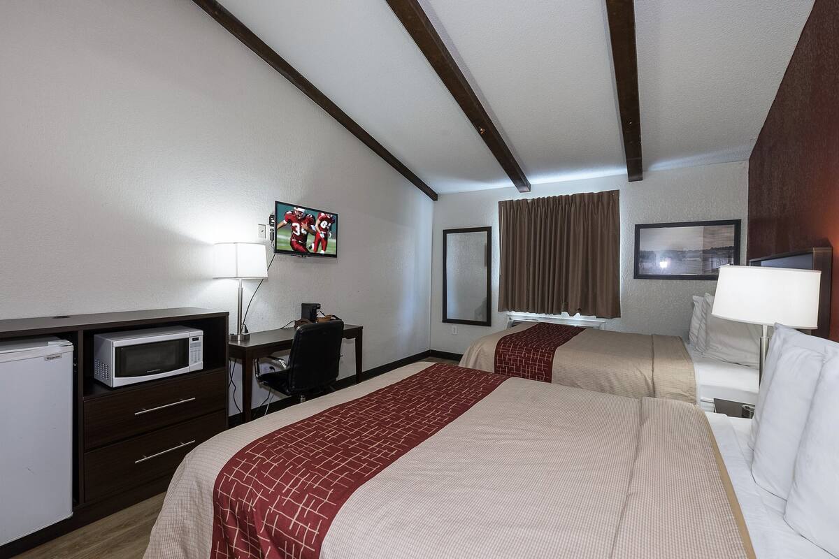 A hotel room with two beds, a microwave, a desk, a chair, a TV, and a window with curtains.