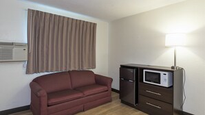 A compact room with a brown leather sofa, a dark wooden cabinet with a microwave, and a wall-mounted air conditioner. - Red Roof Inn Lancaster - Strasburg (Lancaster)
