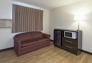 A compact room with a brown leather sofa, a dark wooden cabinet with a microwave, and a wall-mounted air conditioner. - Red Roof Inn Lancaster - Strasburg (Lancaster)