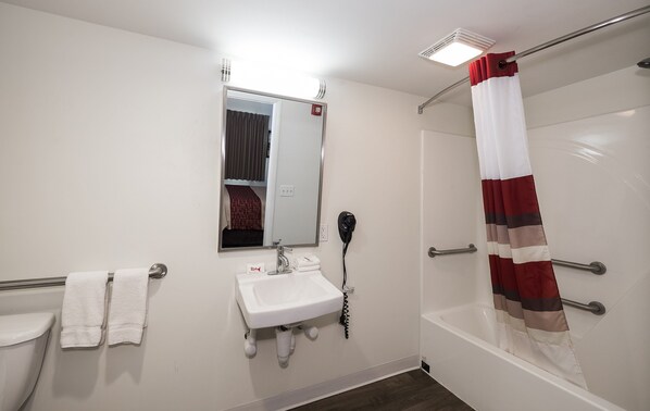 A bathroom with a white sink, a mirror, a towel rack, a shower with a striped curtain, and a handheld hairdryer. - Red Roof Inn Lancaster - Strasburg (Lancaster)