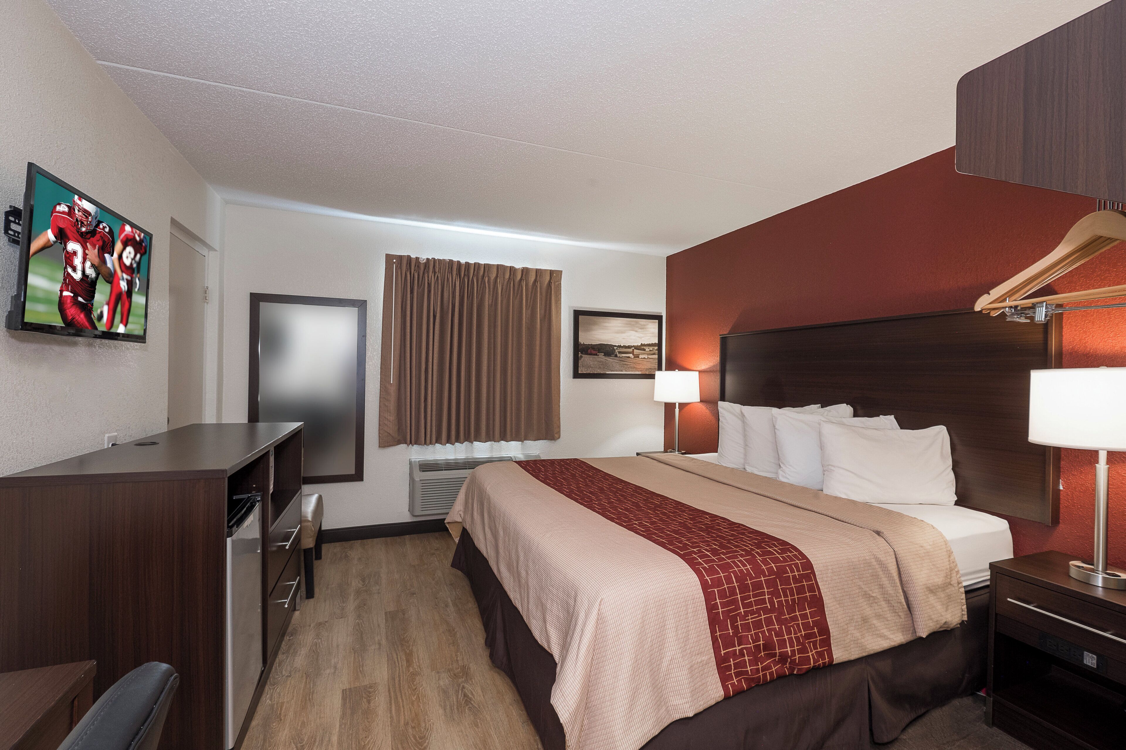 A hotel room with a bed, a desk, a TV, a window with curtains, a wall-mounted picture, and a bedside lamp.