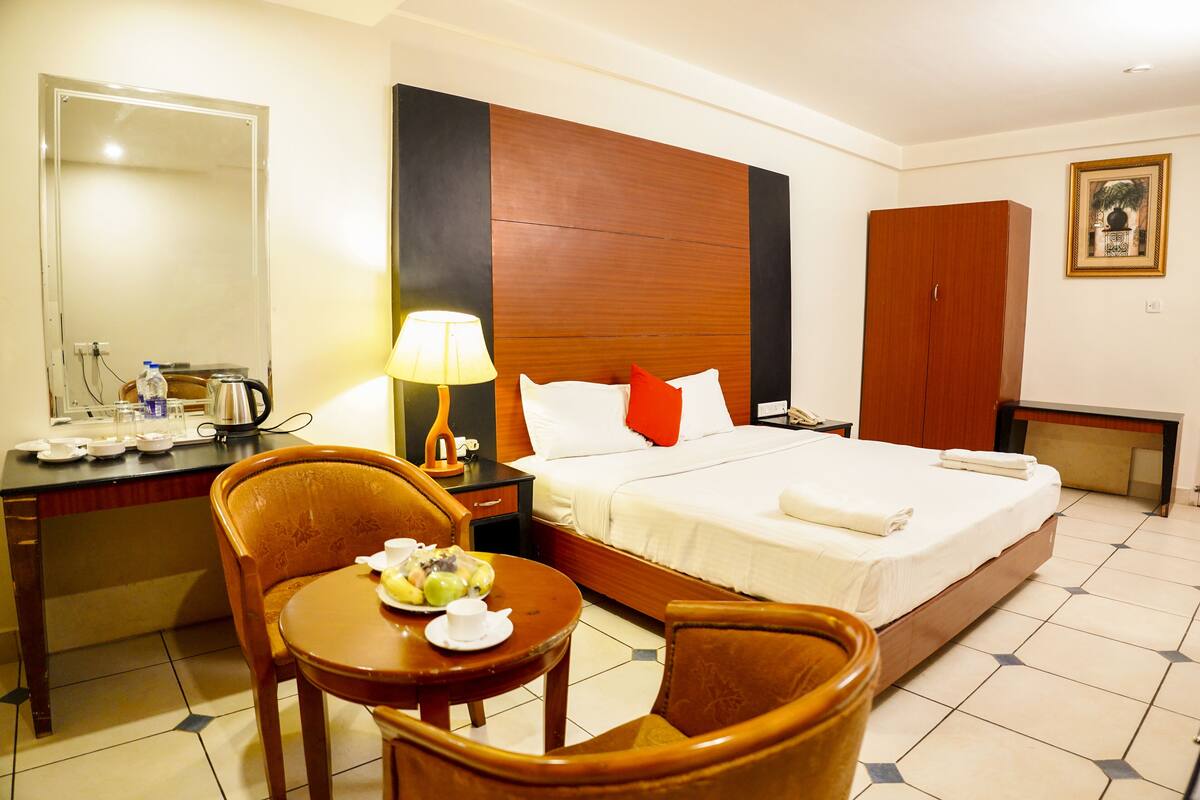 A hotel room with a bed, a desk with a lamp, a chair, and a small table with a plate of food.