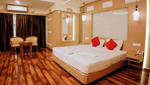 A hotel room with a wooden floor, a bed with white bedding and red pillows, a bedside table with a lamp, a mirror, and a small table with chairs.