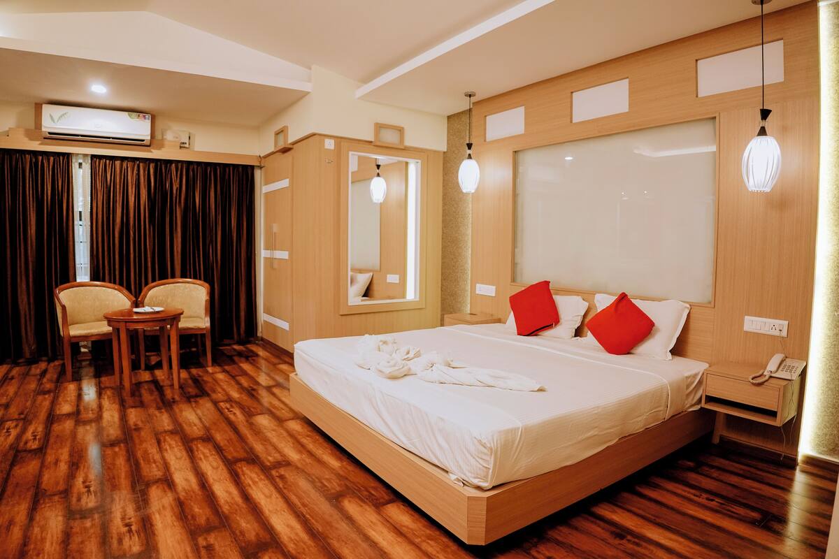 A hotel room with a wooden floor, a bed with white bedding and red pillows, a bedside table with a lamp, a mirror, and a small table with chairs.