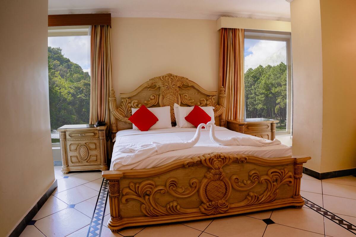 A large bed with ornate headboard and footboard, two red pillows, and a view of trees through the window.