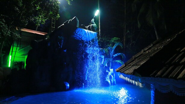 A nighttime waterfall feature with water cascading down a rock formation into a pool, surrounded by tropical vegetation and a traditional-style building.