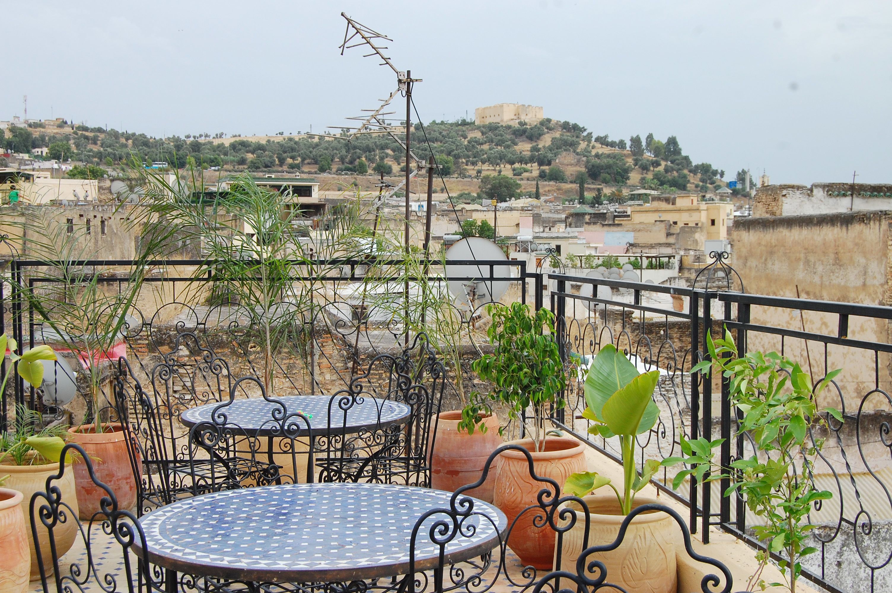 A rooftop terrace with a round metal table and chairs, surrounded by potted plants.