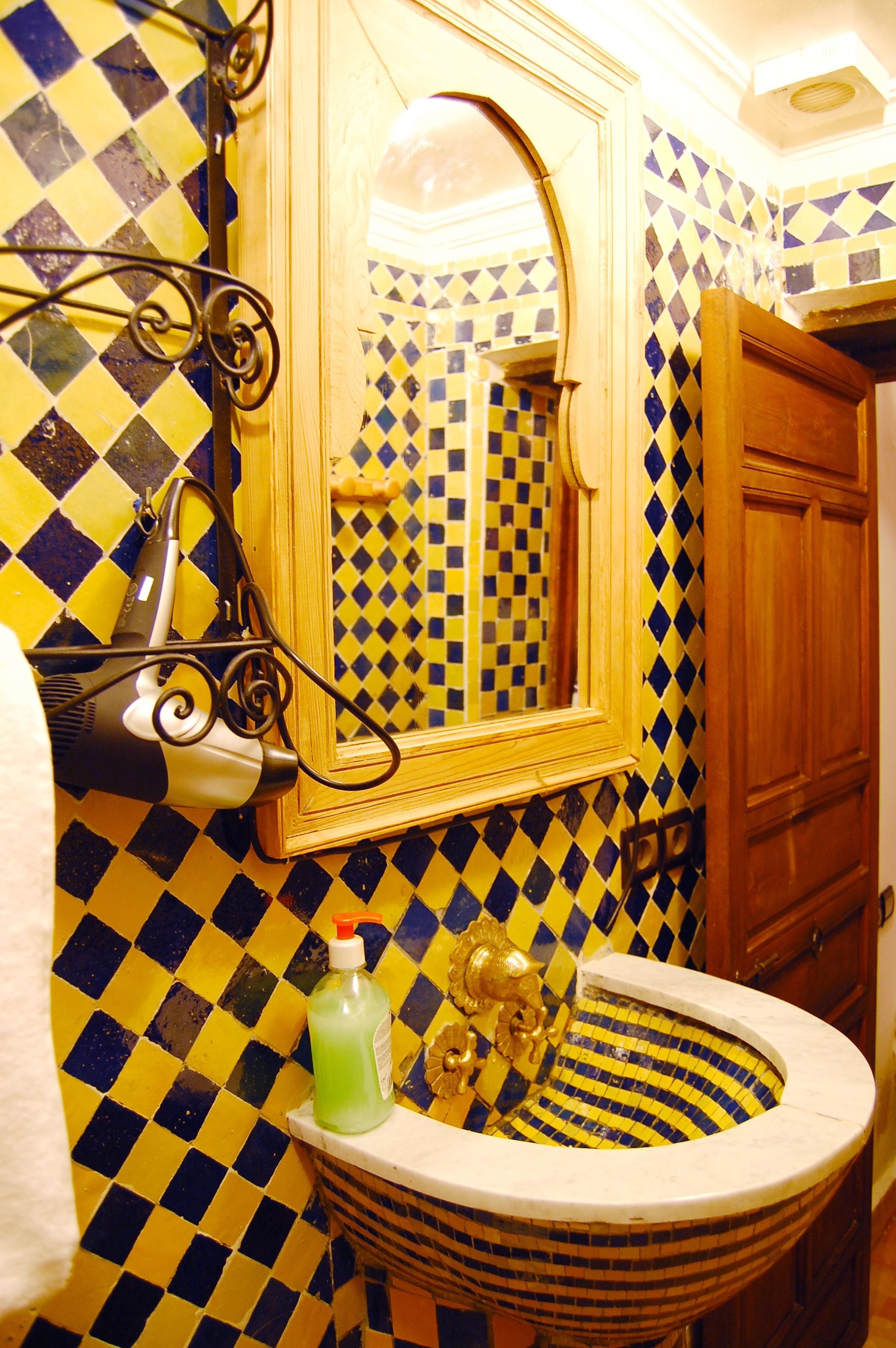 Une salle de bain avec un mur recouvert de carreaux jaunes et bleus formant un motif à carreaux, un miroir et un lavabo agrémenté d’un récipient décoratif.