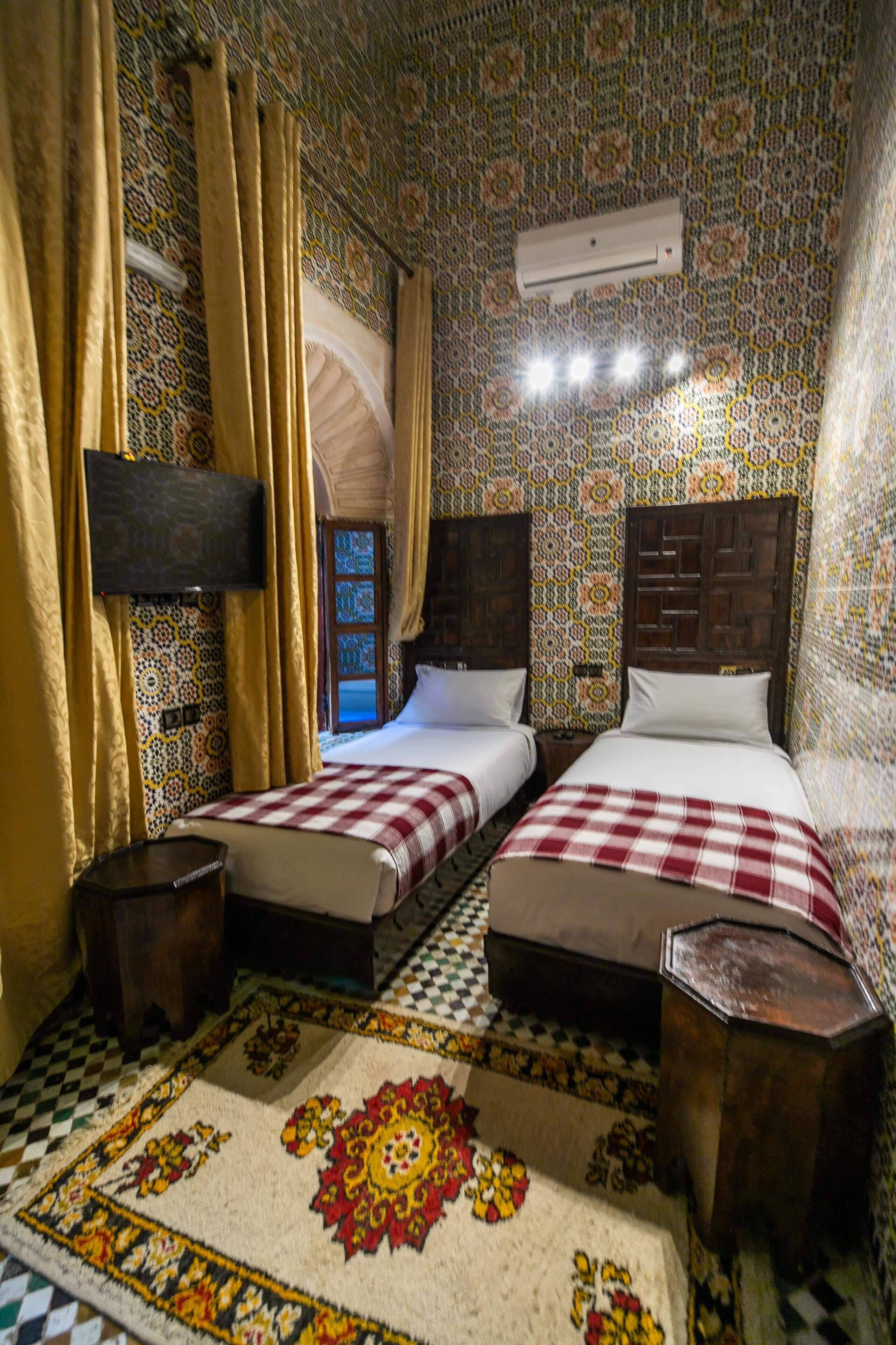 A room with two beds, patterned walls, a checkered rug, and a mounted TV.