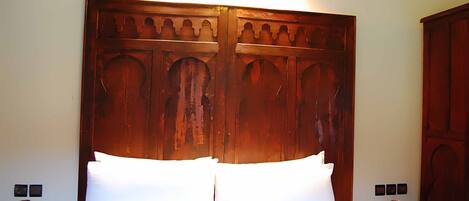 Una cama con ropa de cama blanca, un cabecero de madera con detalles tallados, mesitas de noche con jarrones decorativos y una lámpara de pared.