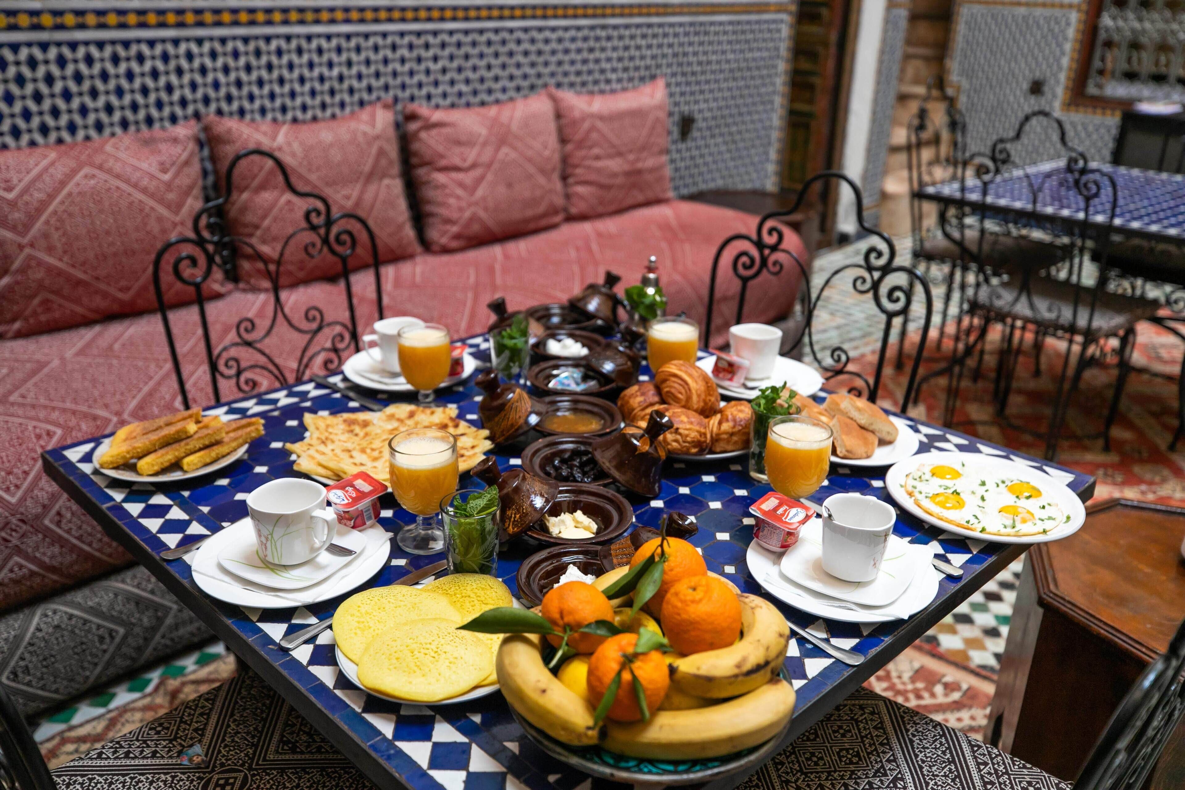 A breakfast table with various foods and drinks, a sofa with cushions, and a patterned wall in the background.