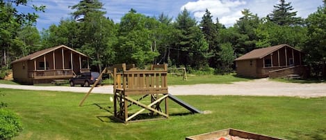 Une structure de jeu en bois et un bac à sable se trouvent dans un espace herbeux, avec deux cabanes en arrière-plan.