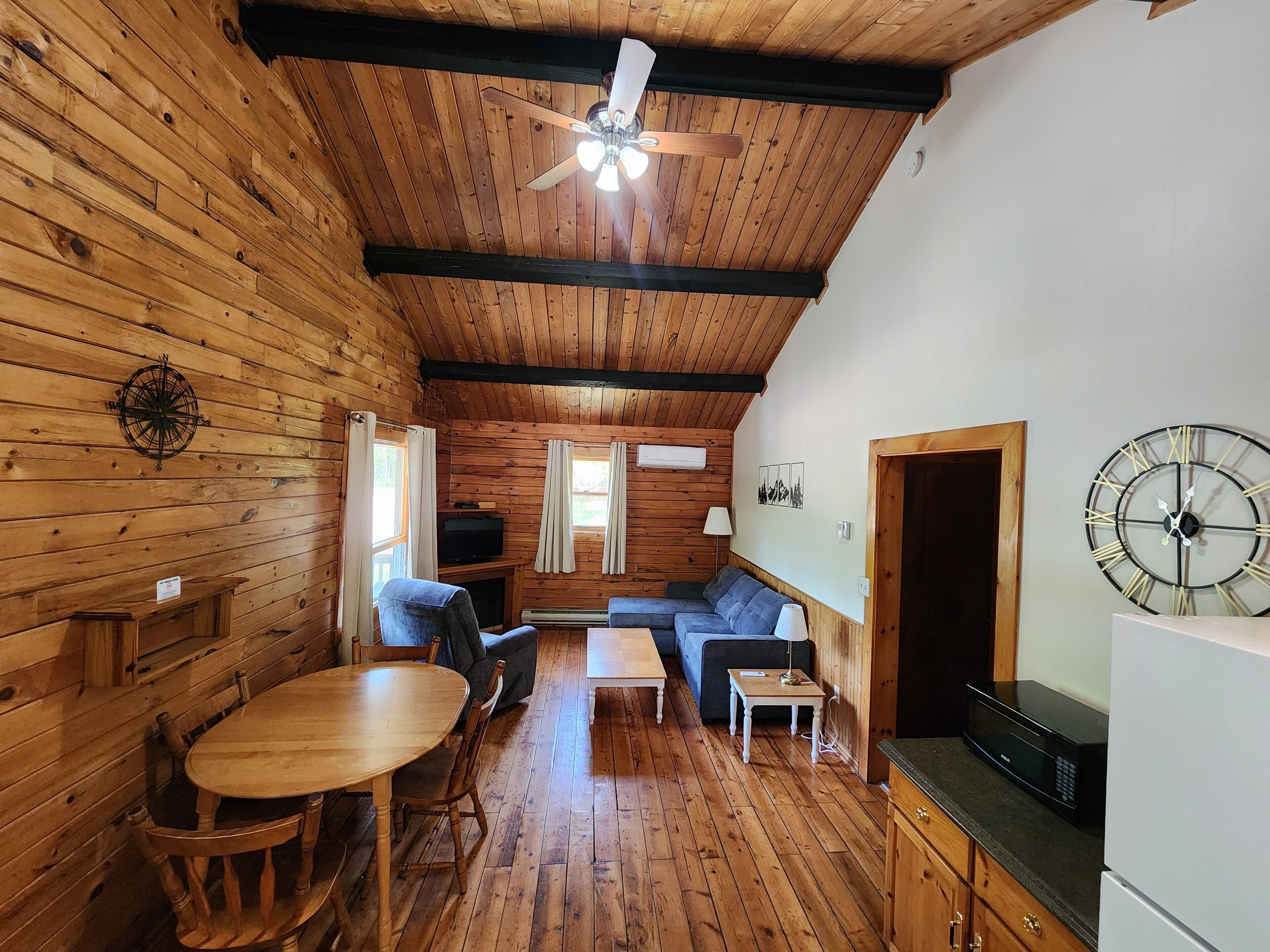 A cozy cabin interior with wooden walls, a ceiling fan, a dining table with chairs, a sofa, a small table, a television, and a clock on the wall.
