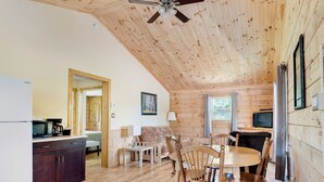 Interior acolhedor de uma cabana, com teto de madeira, área de refeições com mesa redonda e cadeiras, cozinha com geladeira e micro-ondas, e uma janela grande.