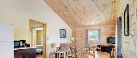 Interior acogedor de una cabaña con techo de madera, un comedor con mesa redonda y sillas, una cocina con refrigerador y microondas, y un ventanal grande.