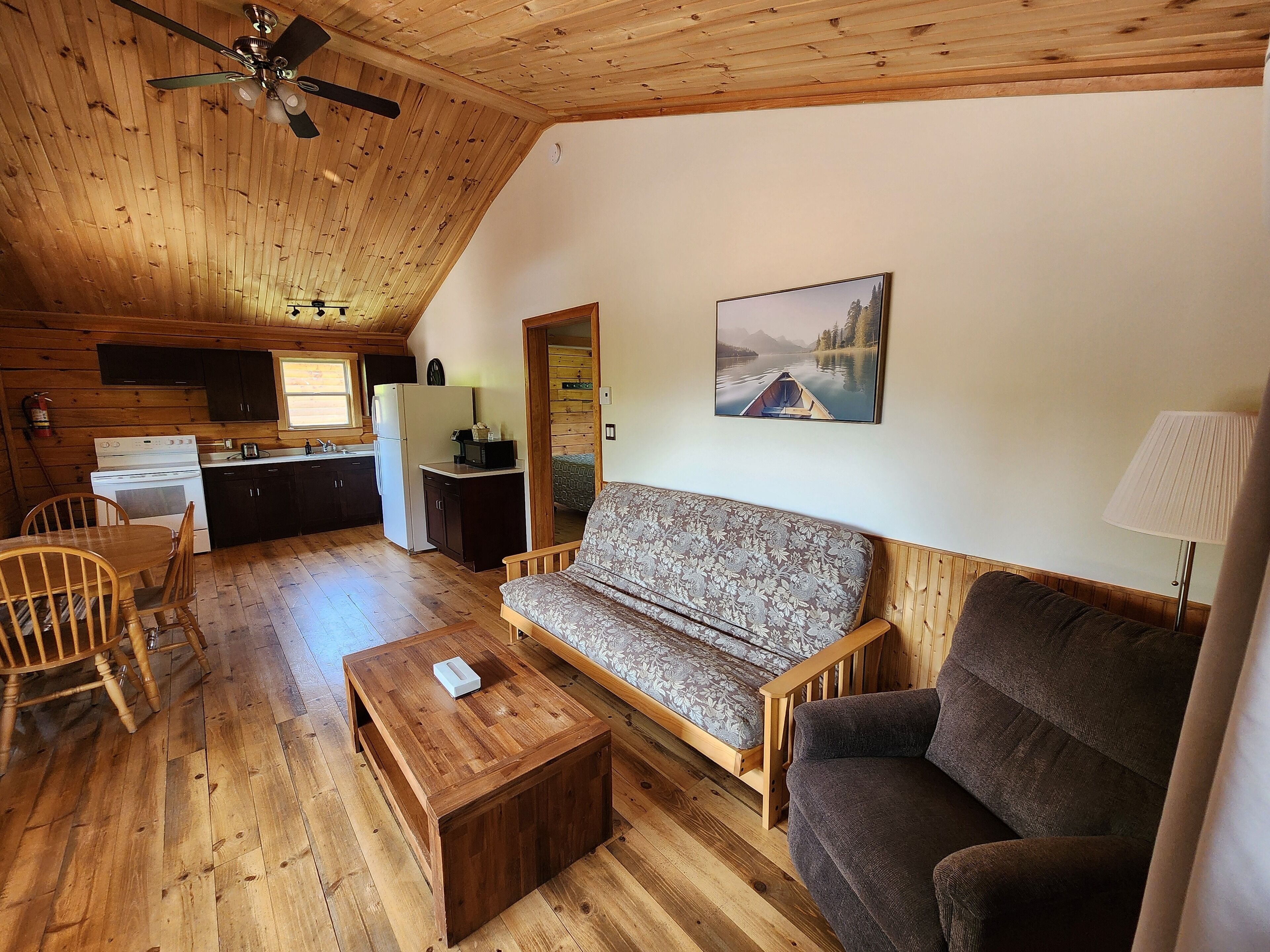 Un interior de cabaña acogedor, con techo de madera, un comedor con mesa y sillas, un sofá, una pequeña mesa de centro de madera, una cocina con refrigerador, estufa y alacenas, y un cuadro en la pared.