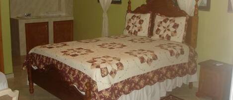 A bedroom with a wooden bed, a bedside table, a nightstand, a chair, a window with curtains, and a patterned rug.