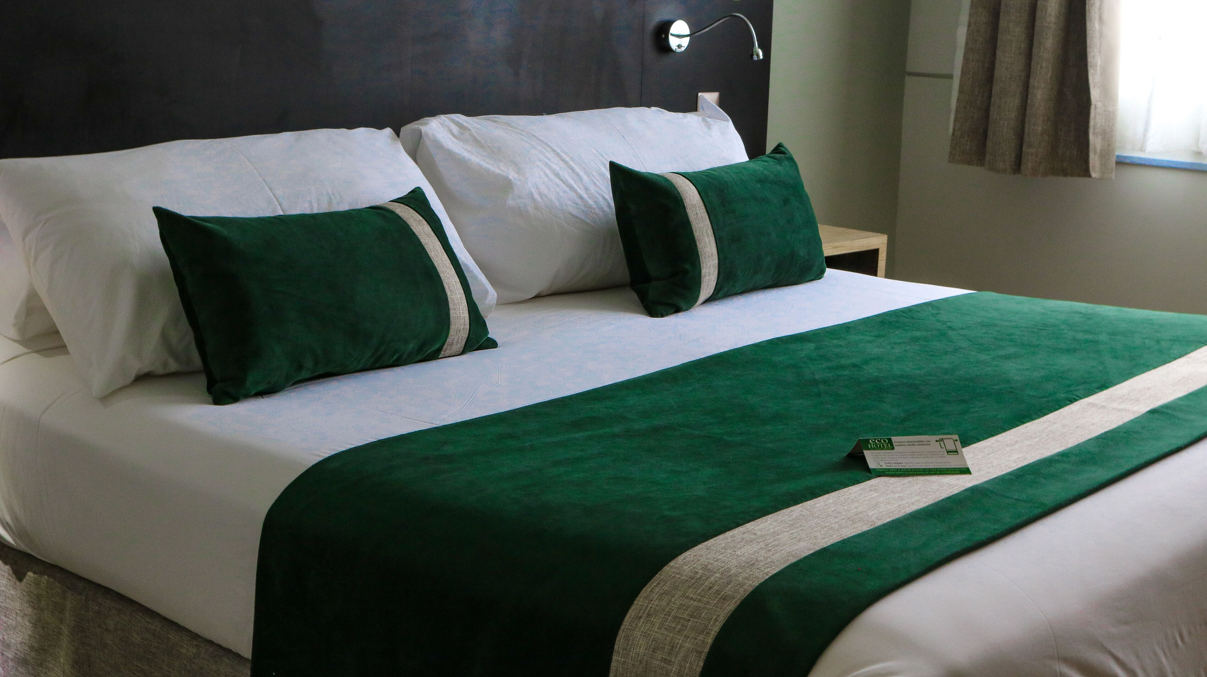 Un lit bien fait, avec une literie blanche et verte, un plaid vert et une petite carte posée sur le lit.