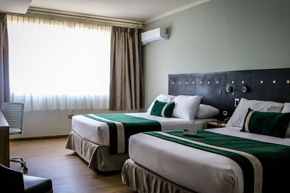 Habitación de hotel con dos camas, un escritorio, una silla y un ventanal con cortinas.