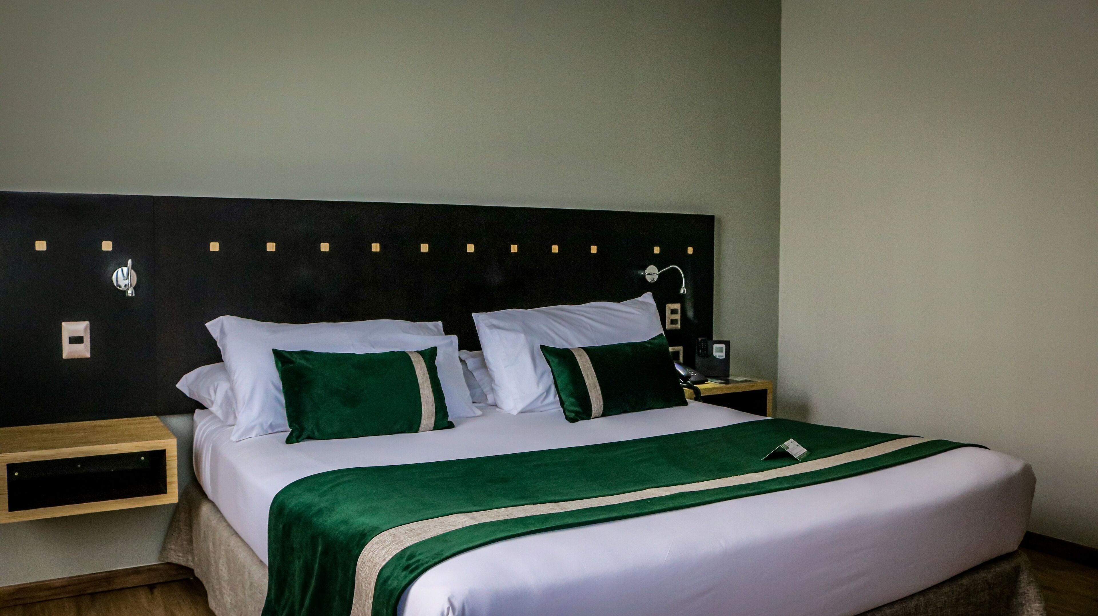 A neatly made bed with white and green bedding, a wooden nightstand with a lamp, and a wall-mounted light fixture.