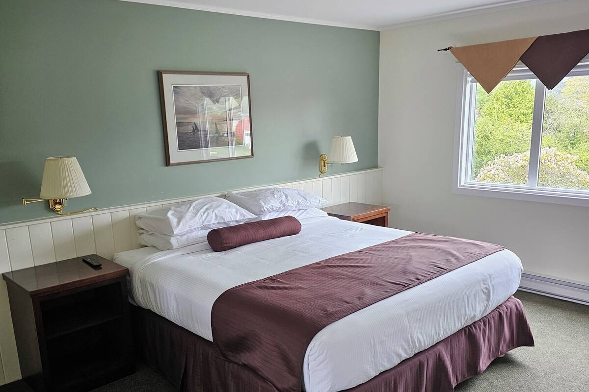 A neatly made bed with a white and maroon bedspread, a single brown pillow, and a framed picture above it. There is a nightstand with a lamp and a remote control on it. A window with a brown awning is visible in the background.