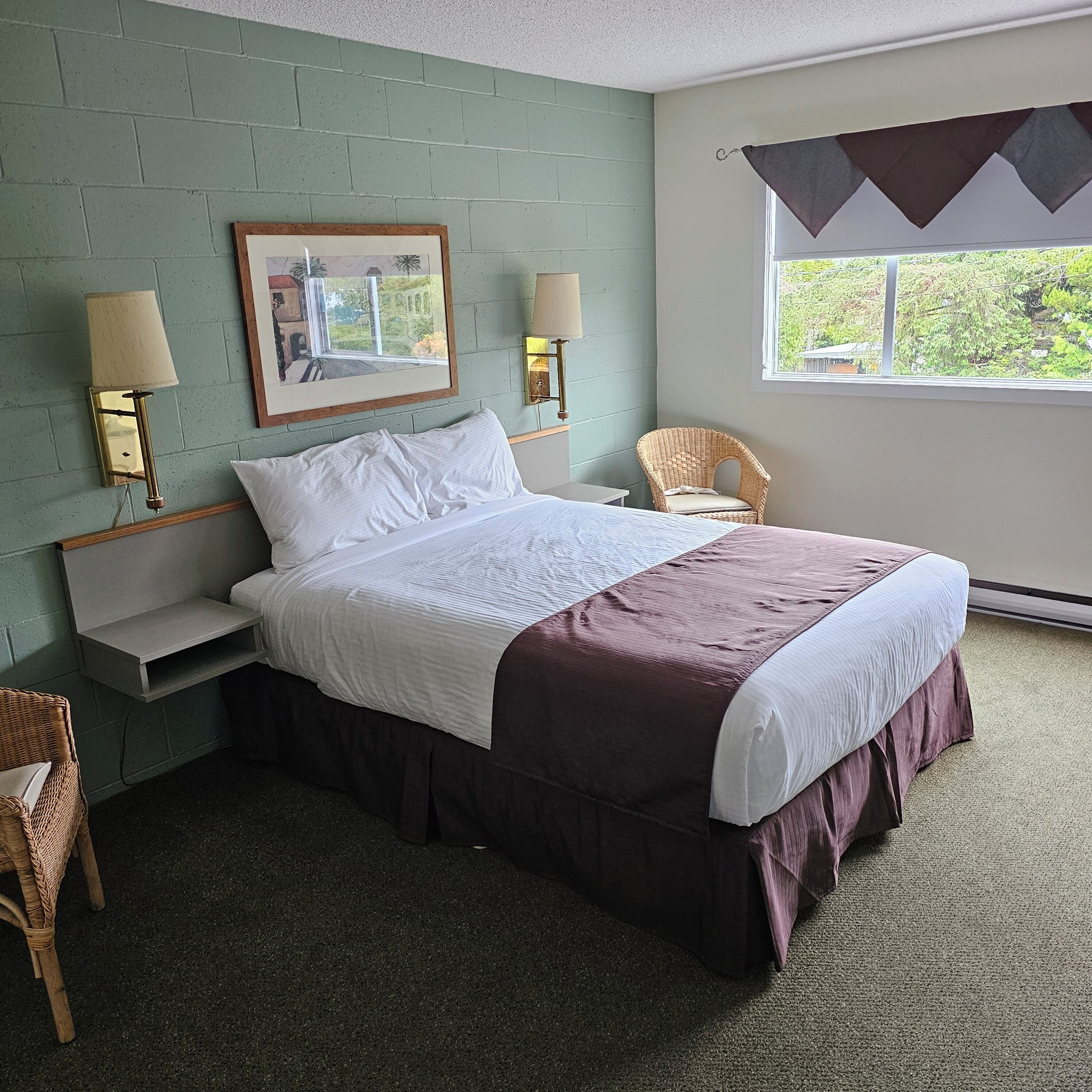 A neatly made bed with a white and brown bedspread, two bedside lamps, a wicker chair, a framed picture on the wall, and a window with a view of greenery.