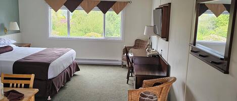 Habitación de hotel con cama, escritorio, silla, televisión y ventana con vista a follaje.