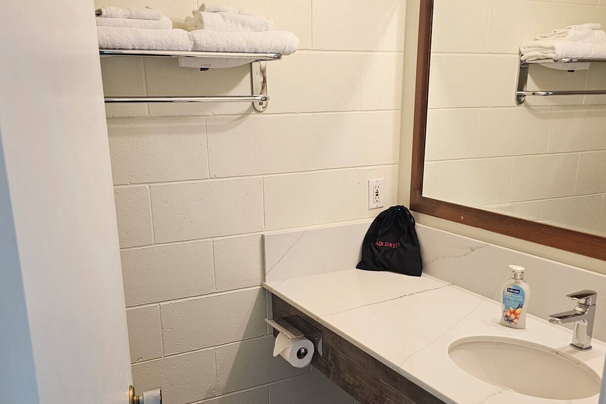 A bathroom with a white sink, a mirror, a towel rack, a trash can, and a black bag on the counter.