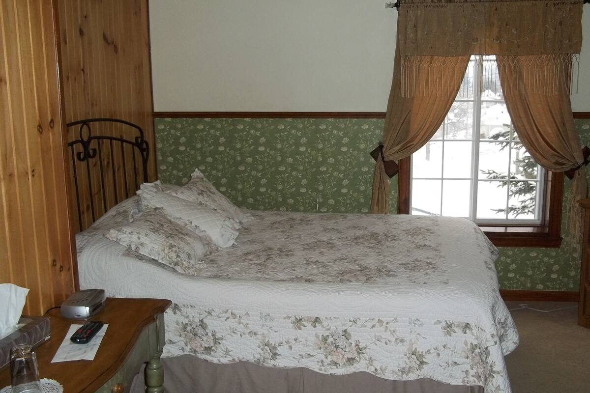 A bedroom with a wooden headboard, a floral bedspread, a bedside table with a lamp, a remote control, and a window with curtains.