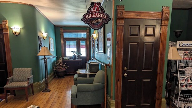 A room with a wooden floor, a green wall, a door, a sign reading 'Rickards,' a lamp, a chair, and a table. - Auberge Le St-Gab (Saint-Gabriel)