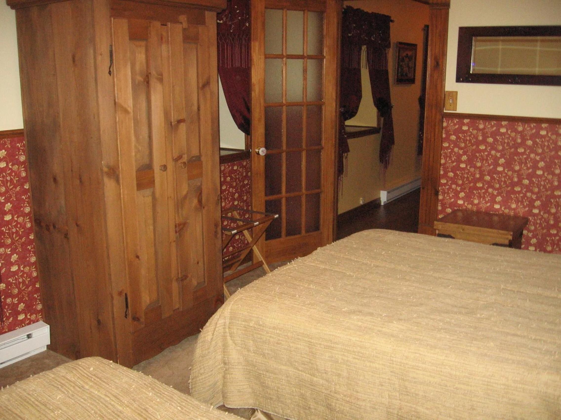 Un dormitorio con un armario de madera, una puerta con paneles de vidrio, una cama con cubrecama color beige y papel tapiz floral.