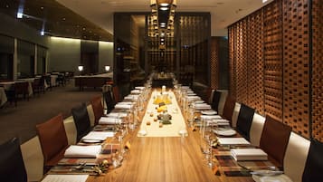 Una larga mesa de comedor dispuesta con múltiples sillas, un arreglo floral central y una pared de ladrillo al fondo.
